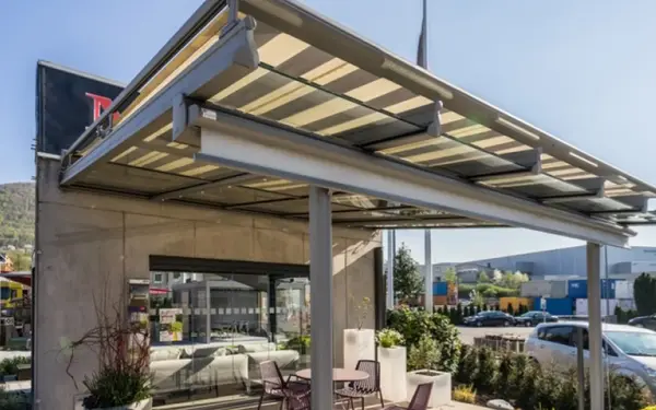 Moderne Terrassenüberdachung aus Glas mit Sonnenschutz und Outdoor-Möbeln vor Gebäude.